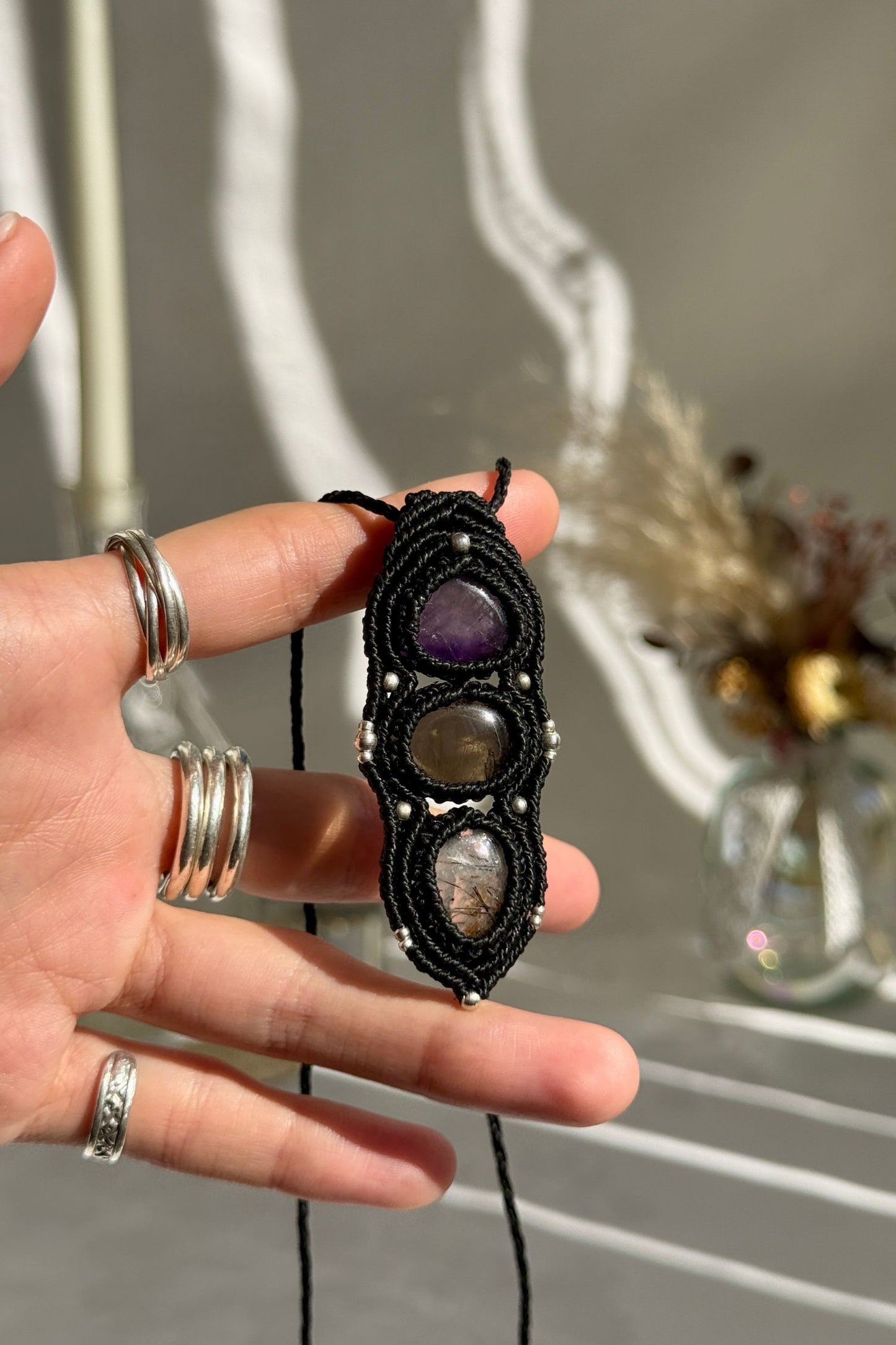 Hand holding a handmade Amethyst, Smoky Quartz and Tourmaline Quartz necklace, highlighting the intricate macrame work and the natural glow of the gemstones.