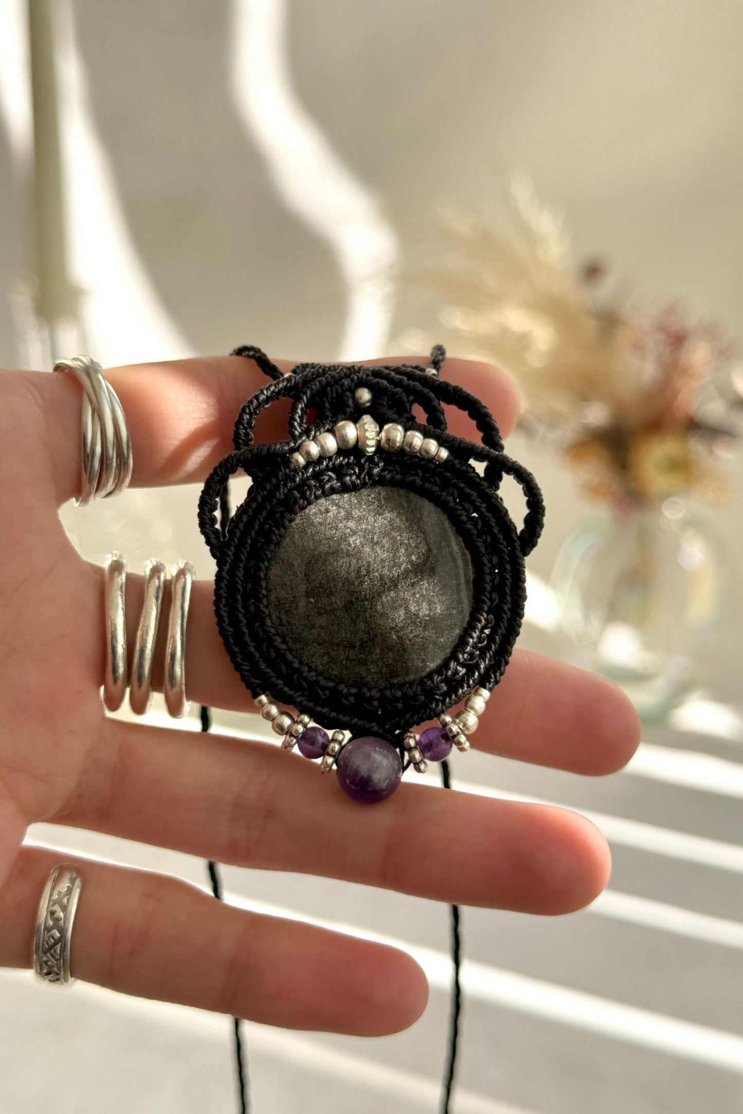 Hand holding a handmade Silver Sheen Obsidian and Amethyst necklace, highlighting the intricate macrame work and the natural glow of the gemstones.