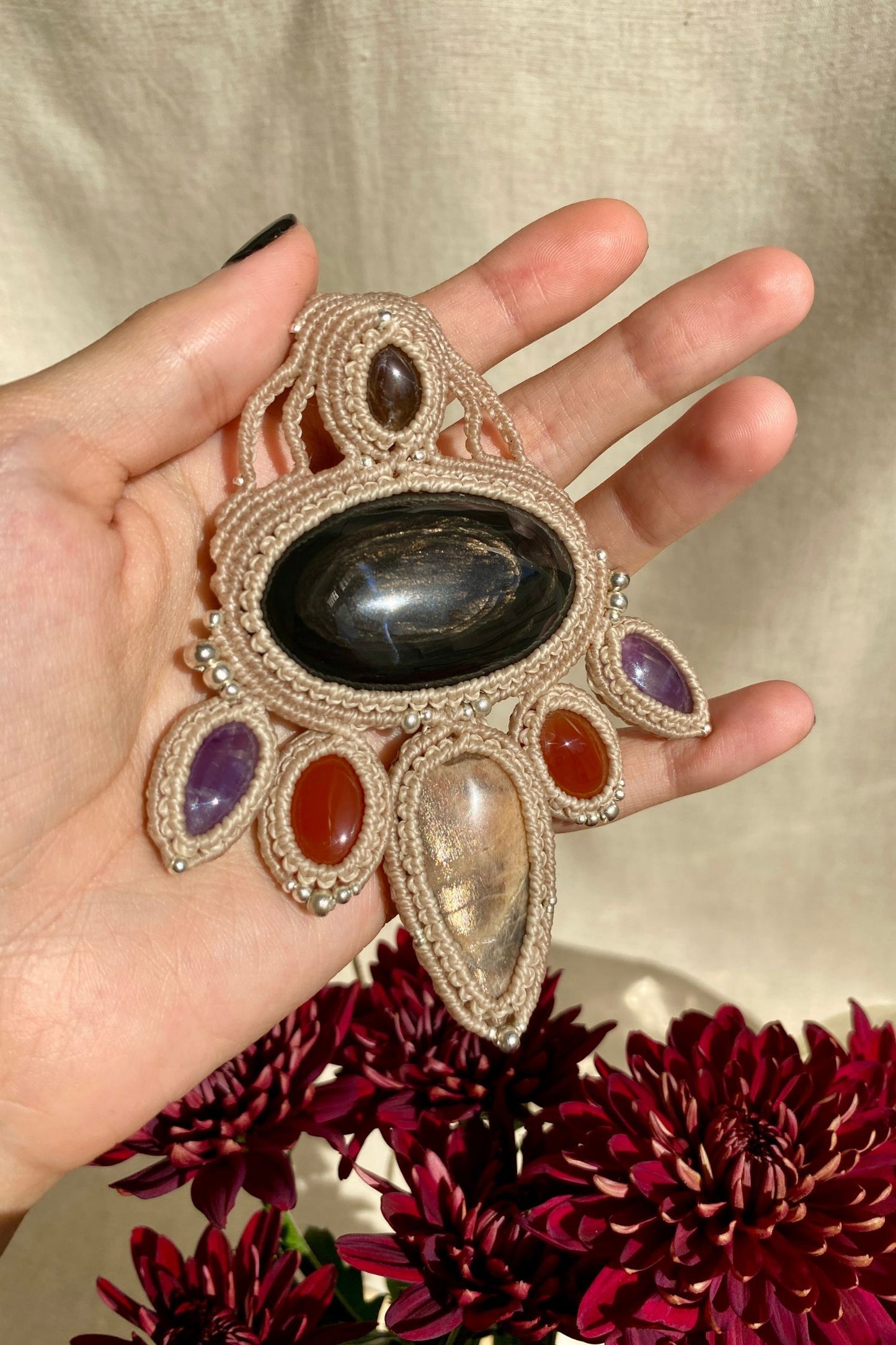 Handmade multi-gemstone macrame necklace held in hand, with sunlight highlighting the bright shine of obsidian, black moonstone, smoky quartz, carnelian, and amethyst gemstones.