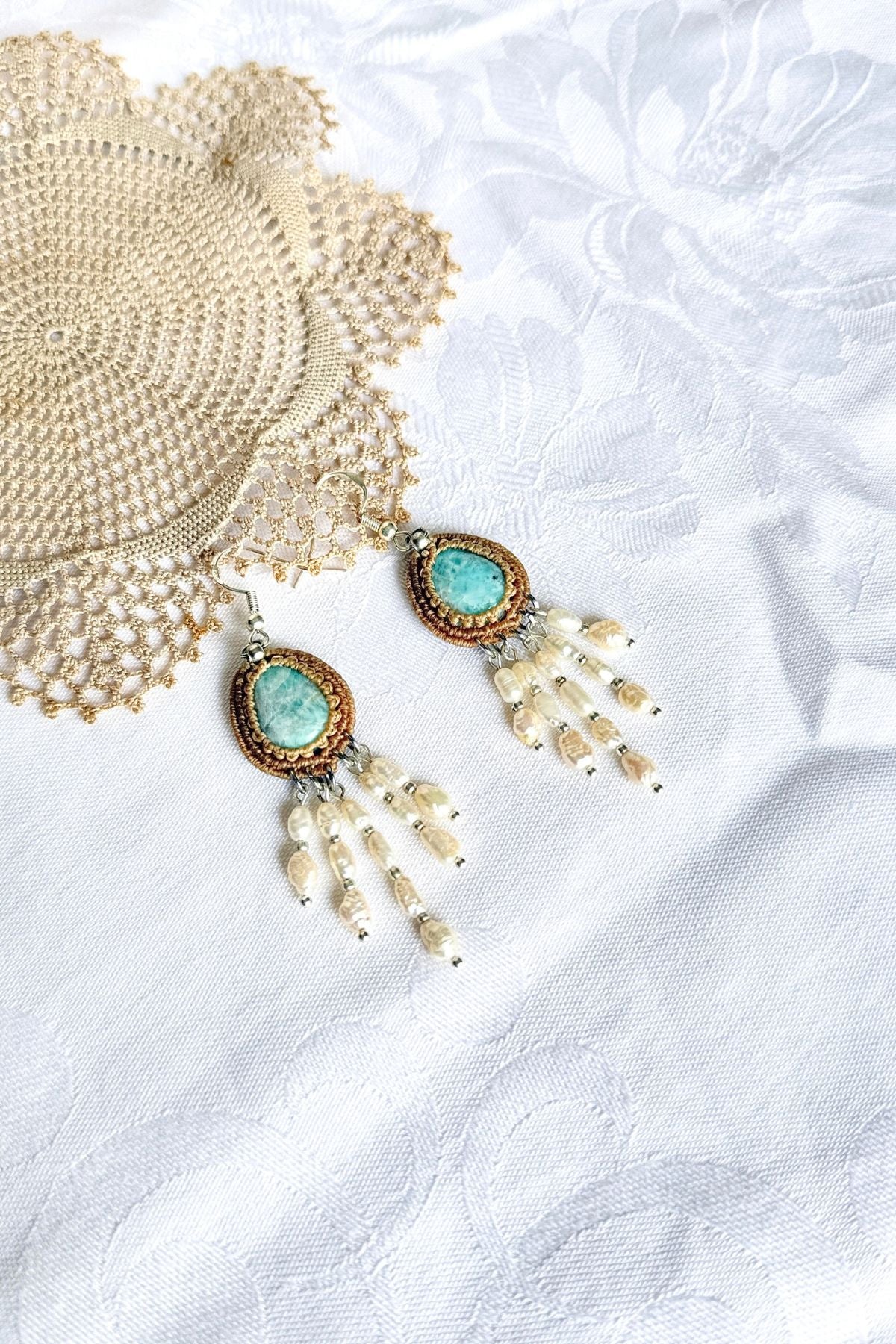 Pair of Larimar macrame earrings with freshwater pearl strands laid on white fabric beside crochet lace.