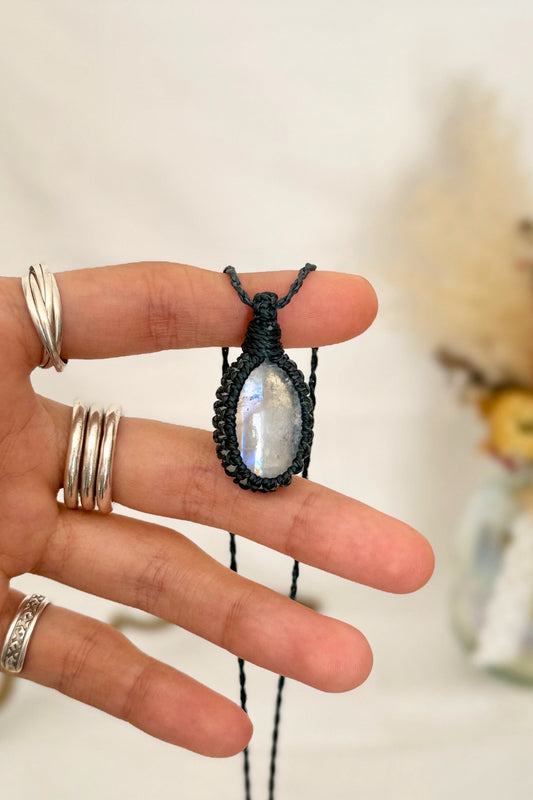 Hand holding a handmade Rainbow Moonstone macrame necklace with a dark blue cord, showcasing the gemstone's mysterious beauty, revealing magical rainbow flashes.