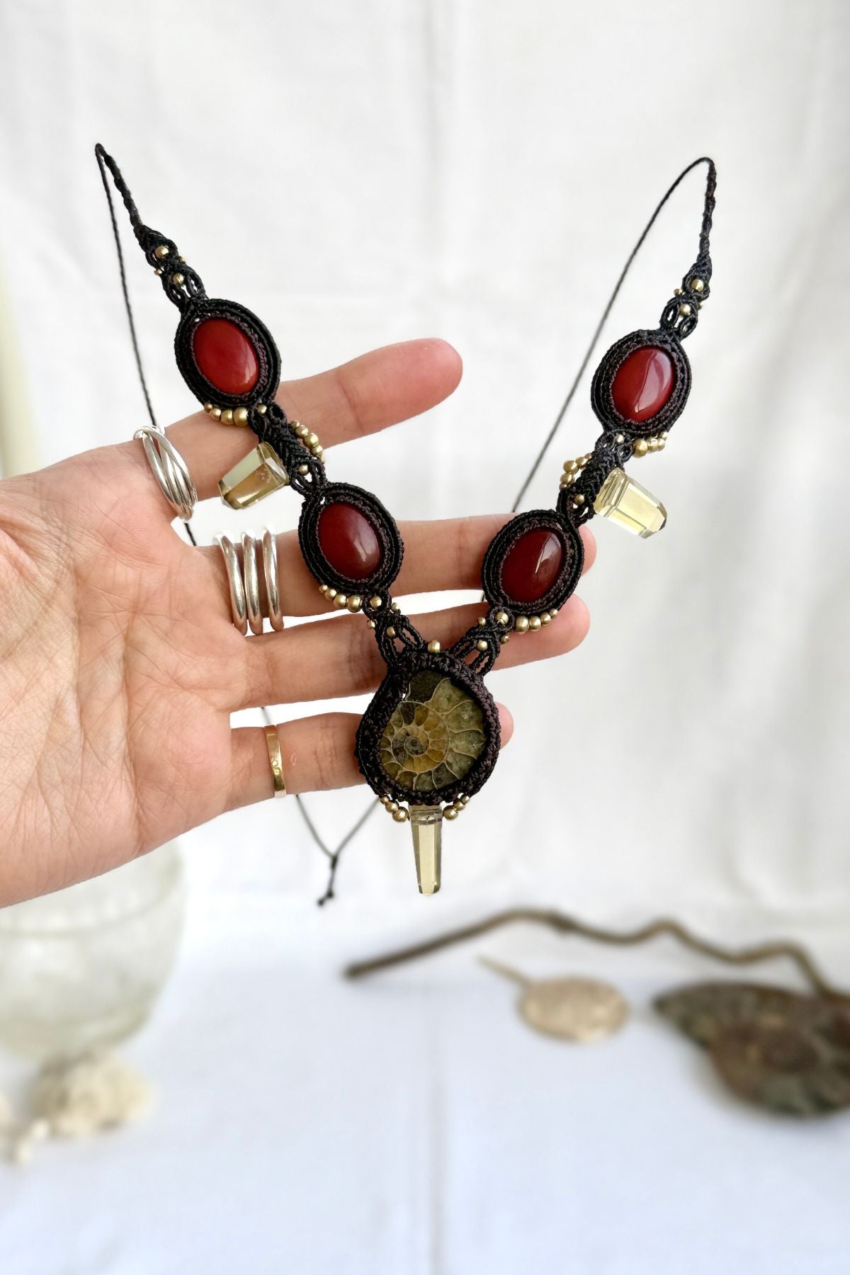 Hand holding V-shaped ammonite necklace showing central yellow ammonite, vivid carnelian stones, smoky quartz points, and macramé craftsmanship
