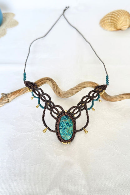 Handmade chrysocolla necklace displayed on white fabric with driftwood, featuring brown macrame and turquoise cord details.