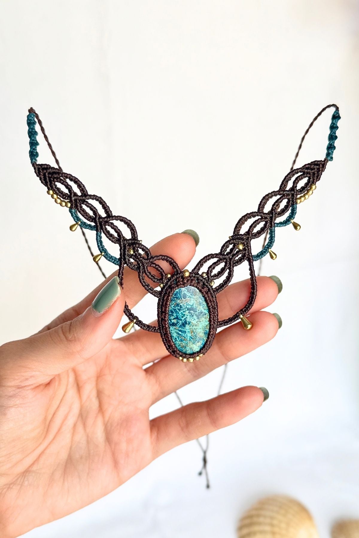 Hand holding a chrysocolla gemstone necklace with brown macrame loops and gold-toned drop accents.