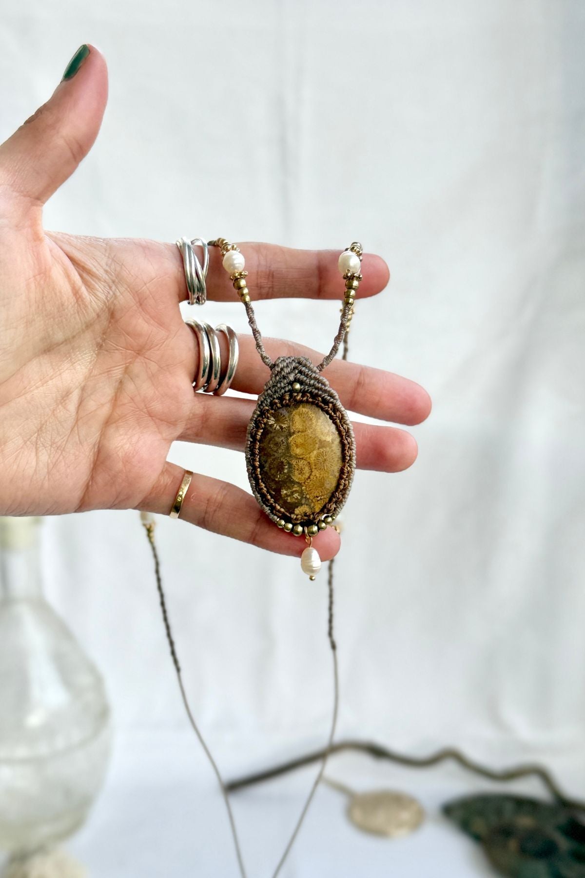 Hand holding fossil coral pendant necklace, showing pendant, pearl drop, and detailed macramé craftsmanship