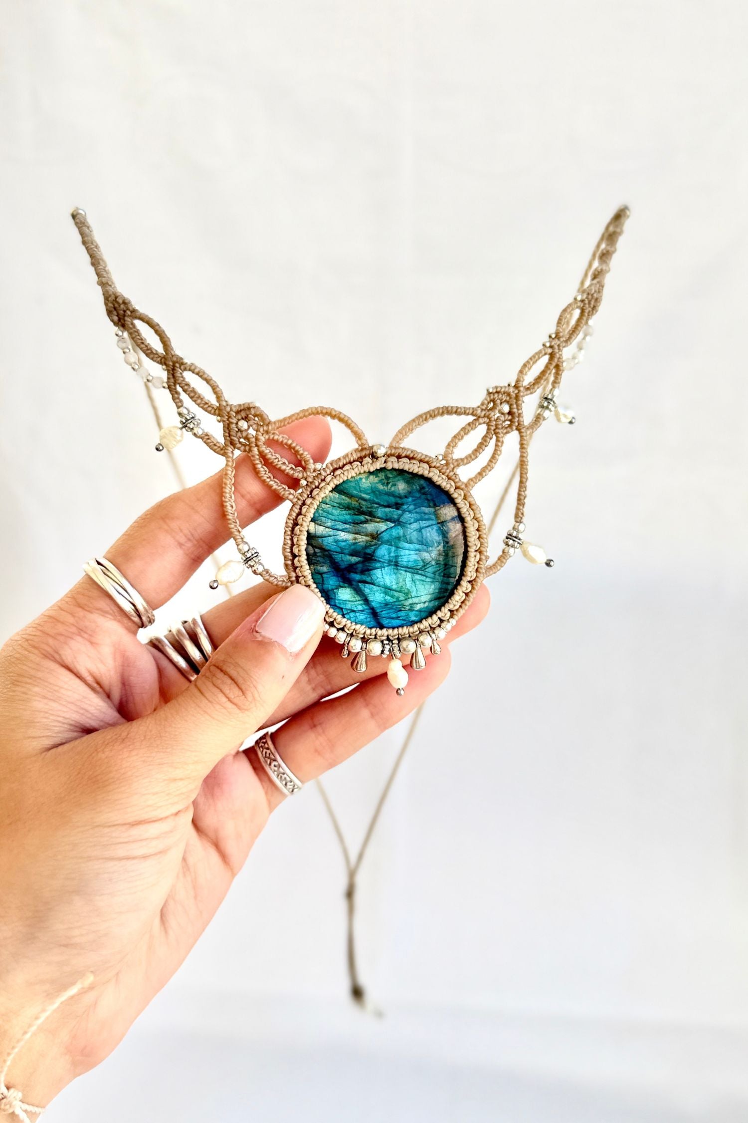 Hand holding a one of a kind Labradorite macrame necklace with deep blue flash, freshwater pearls, silver beads, and beige cord.