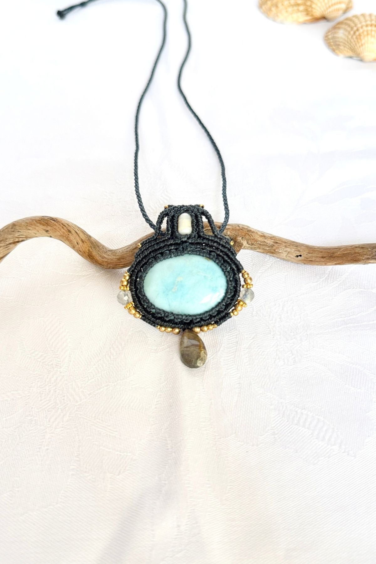 Handmade Larimar necklace in dark blue macrame with a freshwater pearl, Labradorite drop and gold-toned beads, resting on a branch.