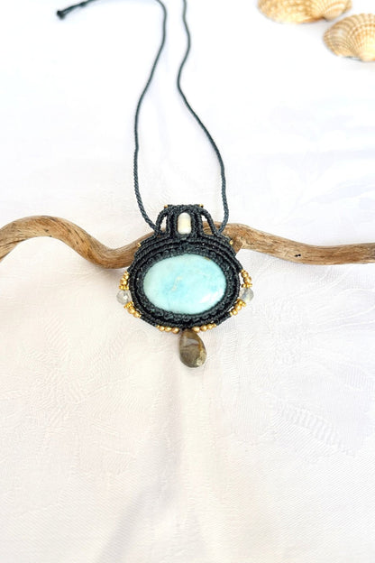 Handmade Larimar necklace in dark blue macrame with a freshwater pearl, Labradorite drop and gold-toned beads, resting on a branch.