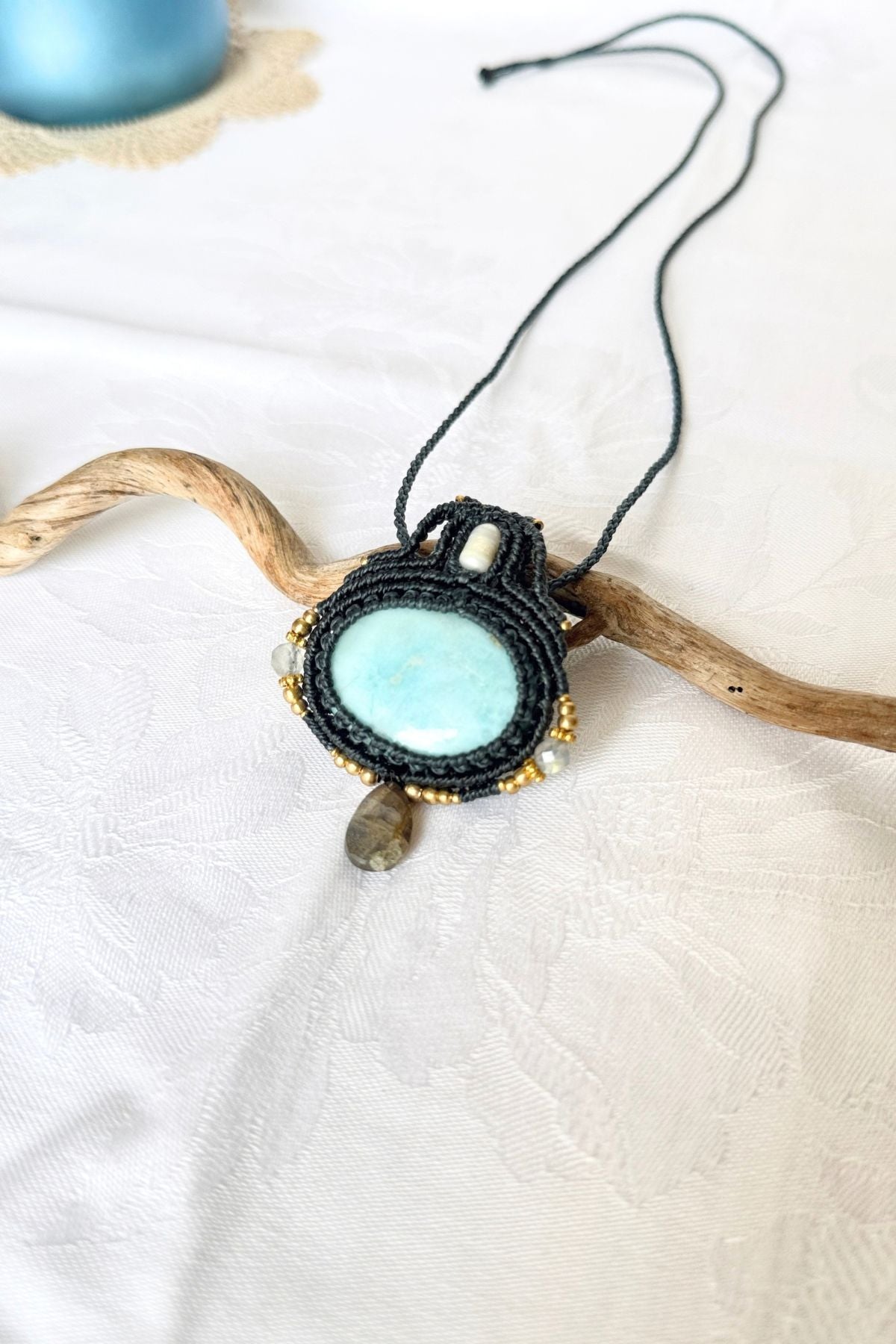 Larimar and Labradorite macrame necklace arranged diagonally on white fabric and a branch, showing the pendant and braided cord.