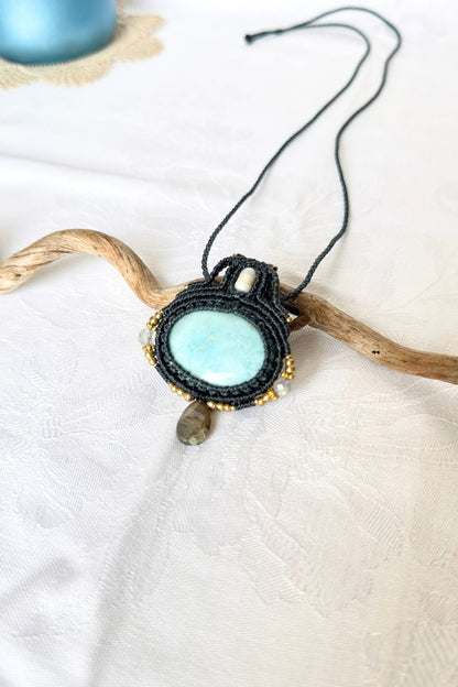 Larimar and Labradorite macrame necklace arranged diagonally on white fabric and a branch, showing the pendant and braided cord.