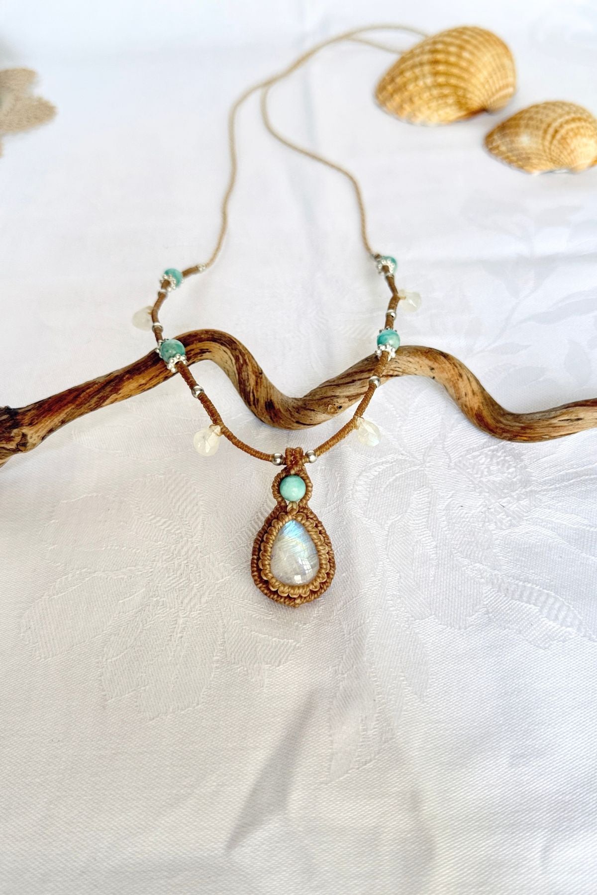 Handmade macrame necklace with a teardrop Rainbow Moonstone pendant and Amazonite beads, displayed over a wooden branch.