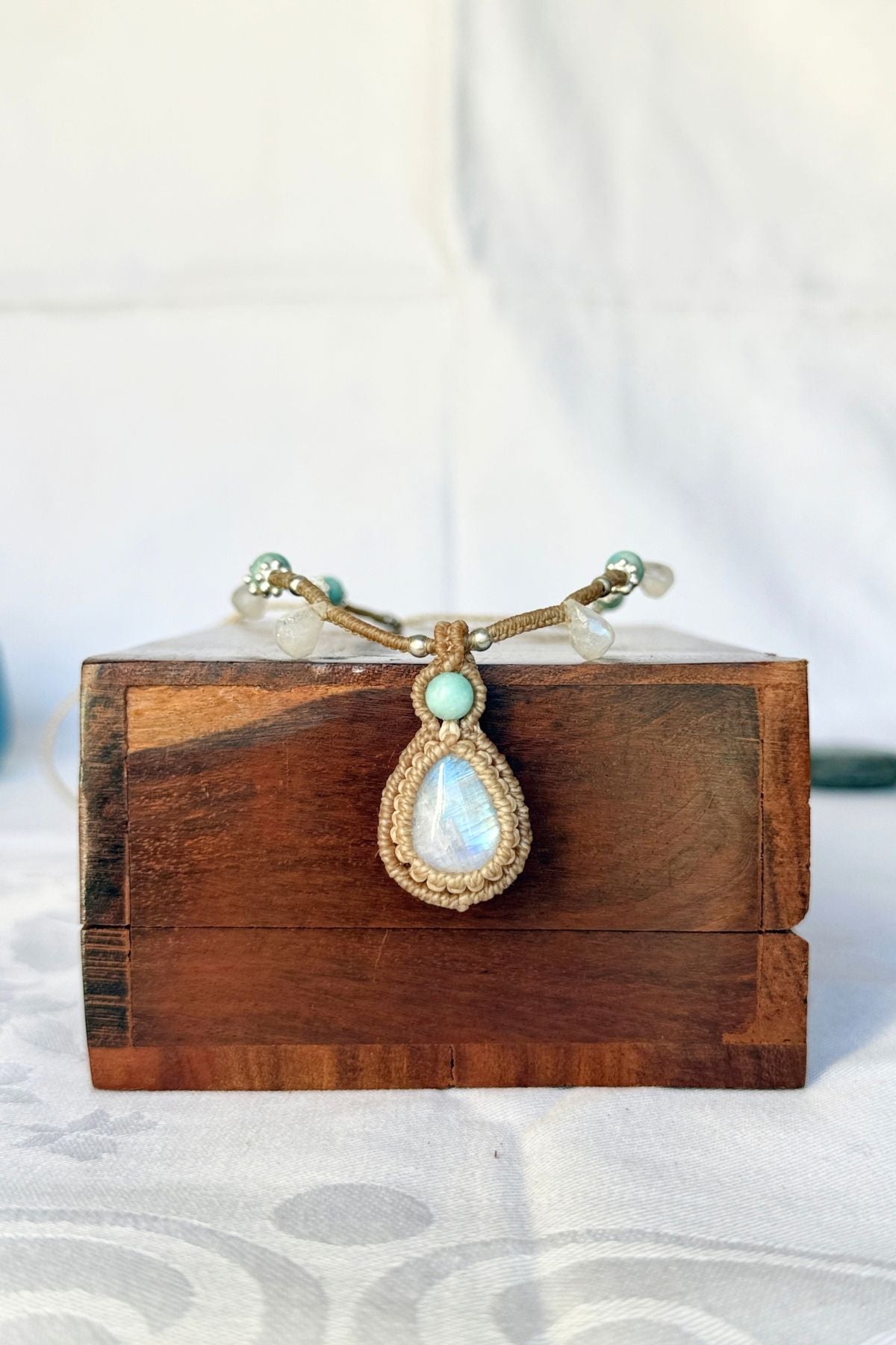 Close-up of a Rainbow Moonstone pendant with a blue flash, framed in tan macrame with an Amazonite accent above.