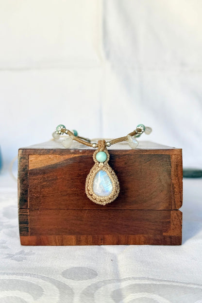 Close-up of a Rainbow Moonstone pendant with a blue flash, framed in tan macrame with an Amazonite accent above.