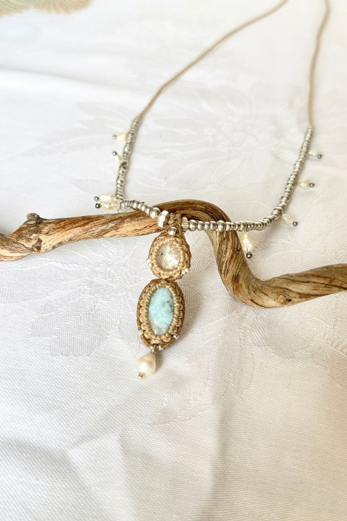 “Angled view of the Moonstone and Larimar pendant, framed in beige macrame with a freshwater pearl drop.