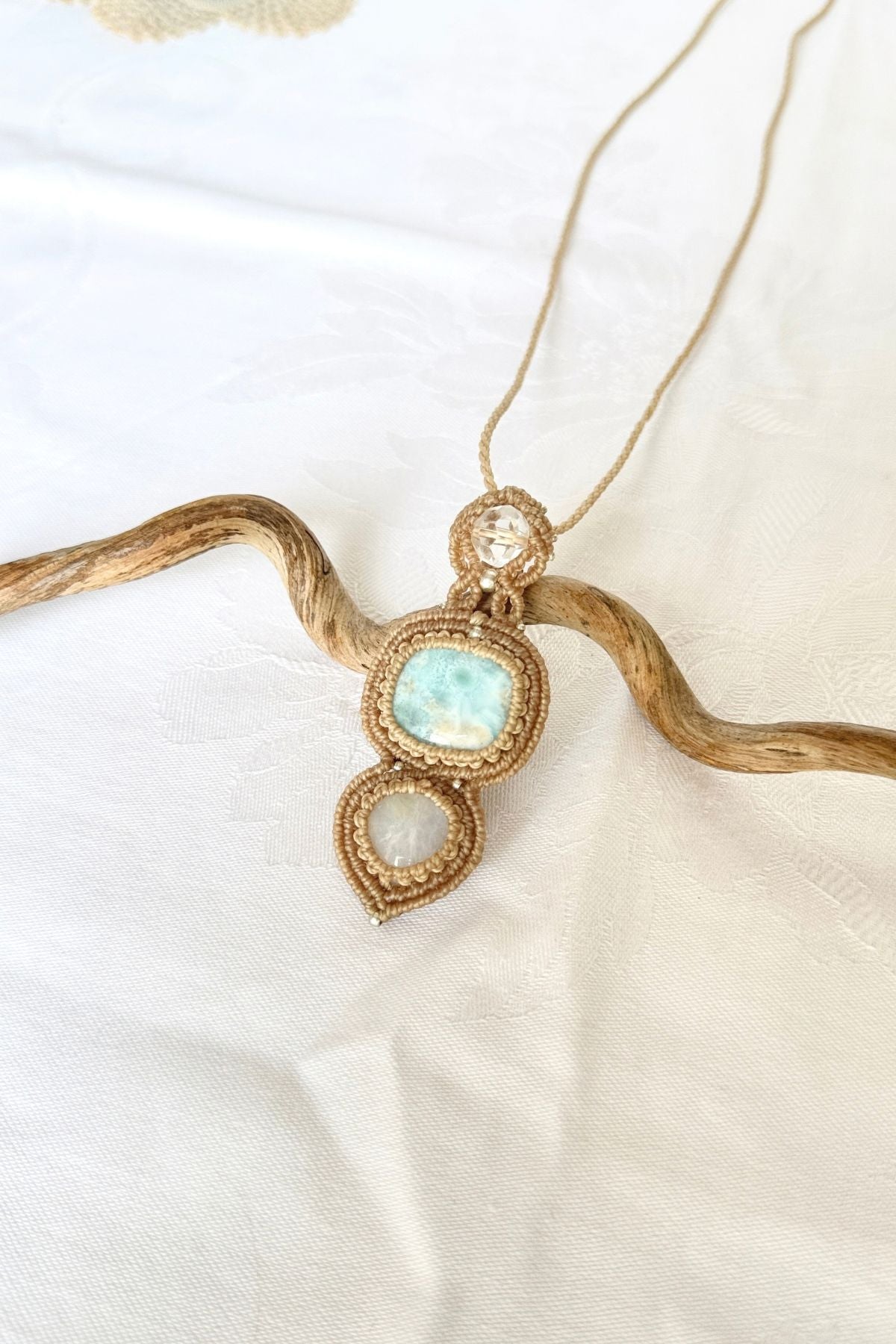 Larimar pendant on white fabric, with clear quartz above and Moonstone below, framed by intricate macrame knots.